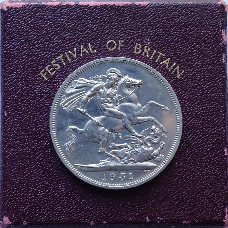 Silver Crown coin of 1951, produced for the Festival of Britain, showing a helmeted and plumed, naked Saint George on horseback, facing right, cloak flying behind him, attacking a dragon with his sword. The date, 1951, is in the exergue, and the coin lies on top of its box, which is dark red, and the text, Festival of Britain, is visible.