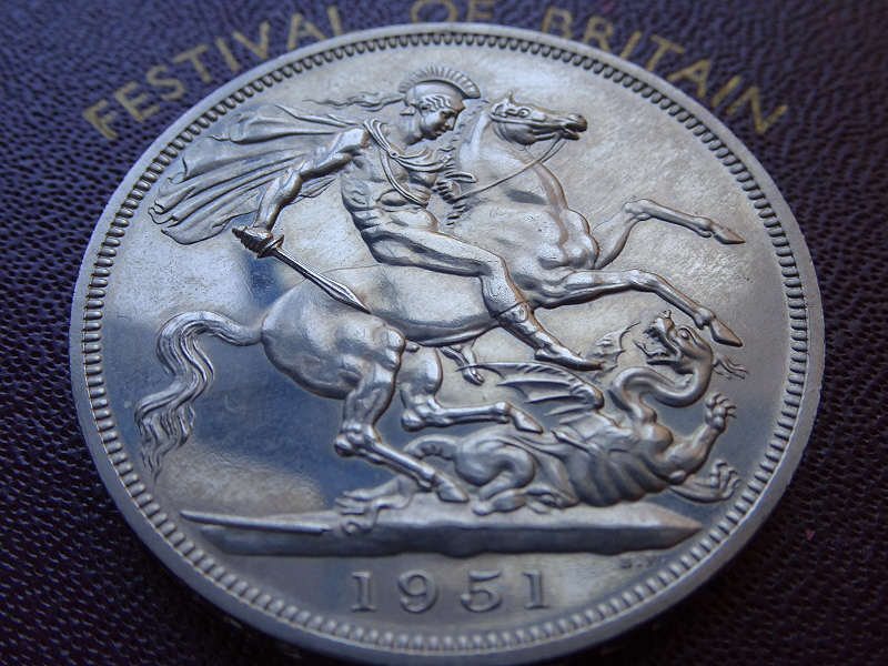 Silver Crown coin of 1951, produced for the Festival of Britain, showing a helmeted and plumed, naked Saint George on horseback, facing right, cloak flying behind him, attacking a dragon with his sword. The date, 1951, is in the exergue, and the coin lies on top of its box, which is dark red, and the text, Festival of Britain, is visible.