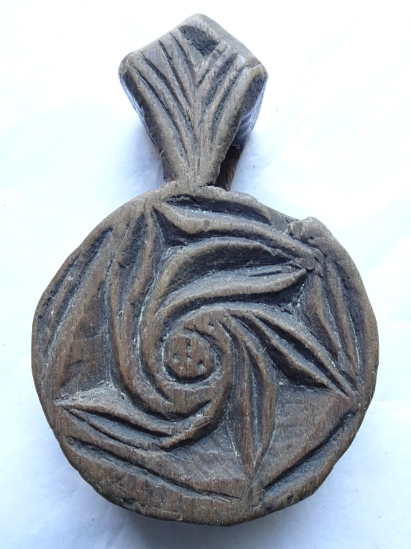 Carved wooden amulet, round with roughly triangular top piece with a hole for a cord to hang it around an animal's neck. The front has a swirly pattern.