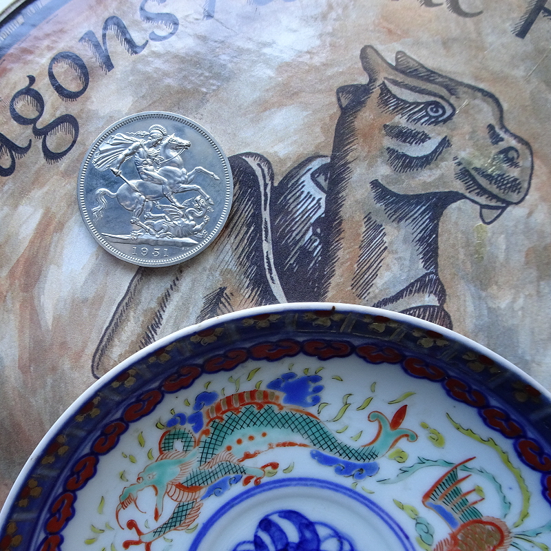 A 1951 Festival of Britain Crown showing Saint George and the Dragon and a Japanese saucer showing a Dragon, on top of a display item from the 2015 Norwich Go-Go-Dragons Festival.