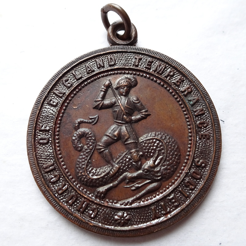 Church of England Temperance Society medal in bronze, showing Saint George attacking a Dragon with a lance (left) and (right) a sceptre across an open book, with a crown at the top. The text on the book reads, "Whether therefore you eat or drink or what so ever ye do do all to the glory of God".